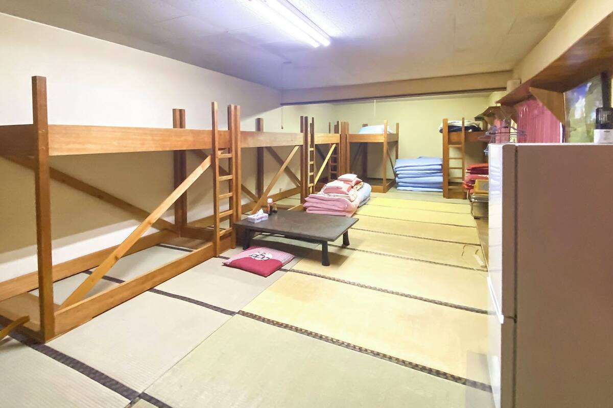Een kamer met tatami vloerbedekking, houten stapelbedden, een kleine tafel met een kussen en een koelkast.