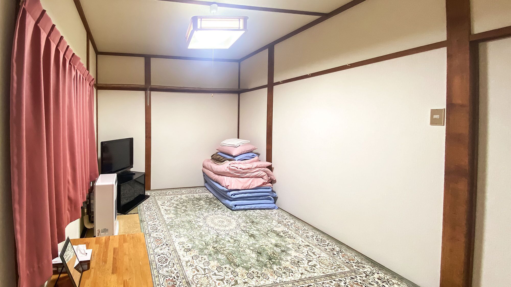 Una habitación con televisión, ropa de cama doblada, un tapete estampado y una ventana con marco de madera.