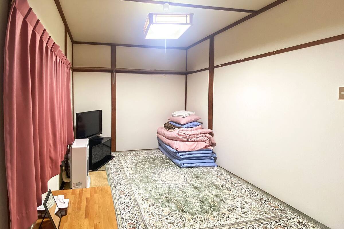 Een kamer met een televisie, een stapel opgevouwen beddengoed, een kamerkleed met een patroon en een raam met houten kader.