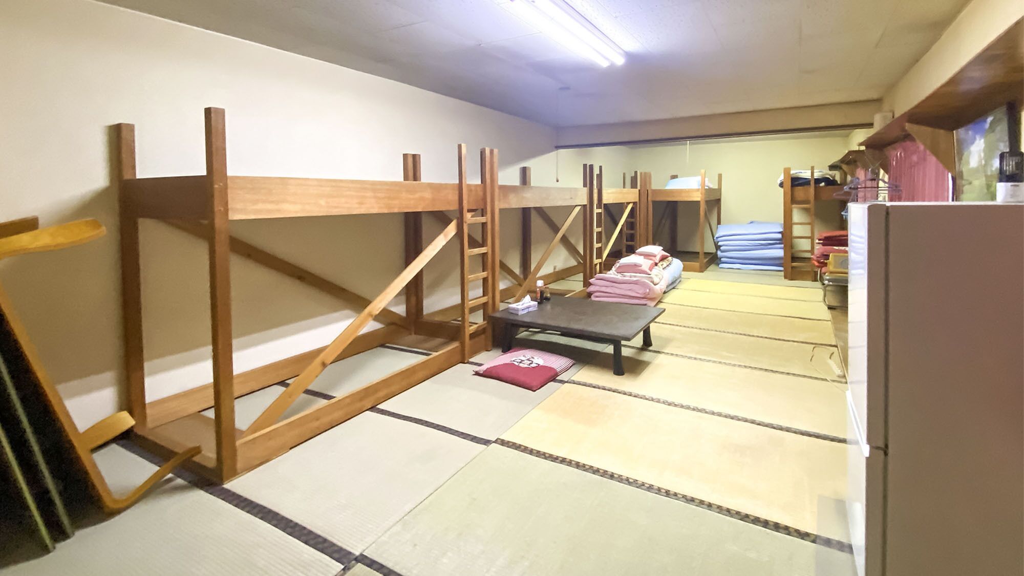 Una habitación con suelo de tatami, literas de madera, una pequeña mesa con un cojín y un refrigerador.