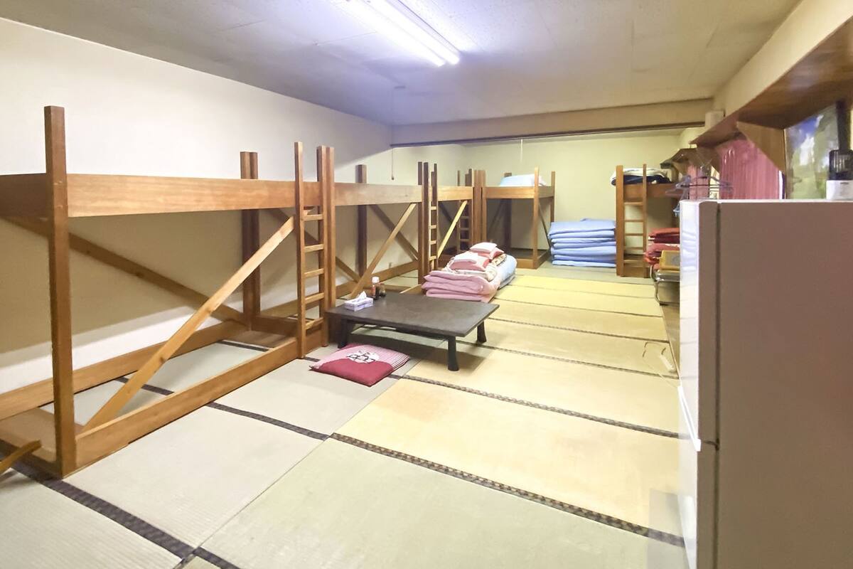 Een kamer met tatami vloerbedekking, houten stapelbedden, een klein tafeltje met een kussen en een koelkast.