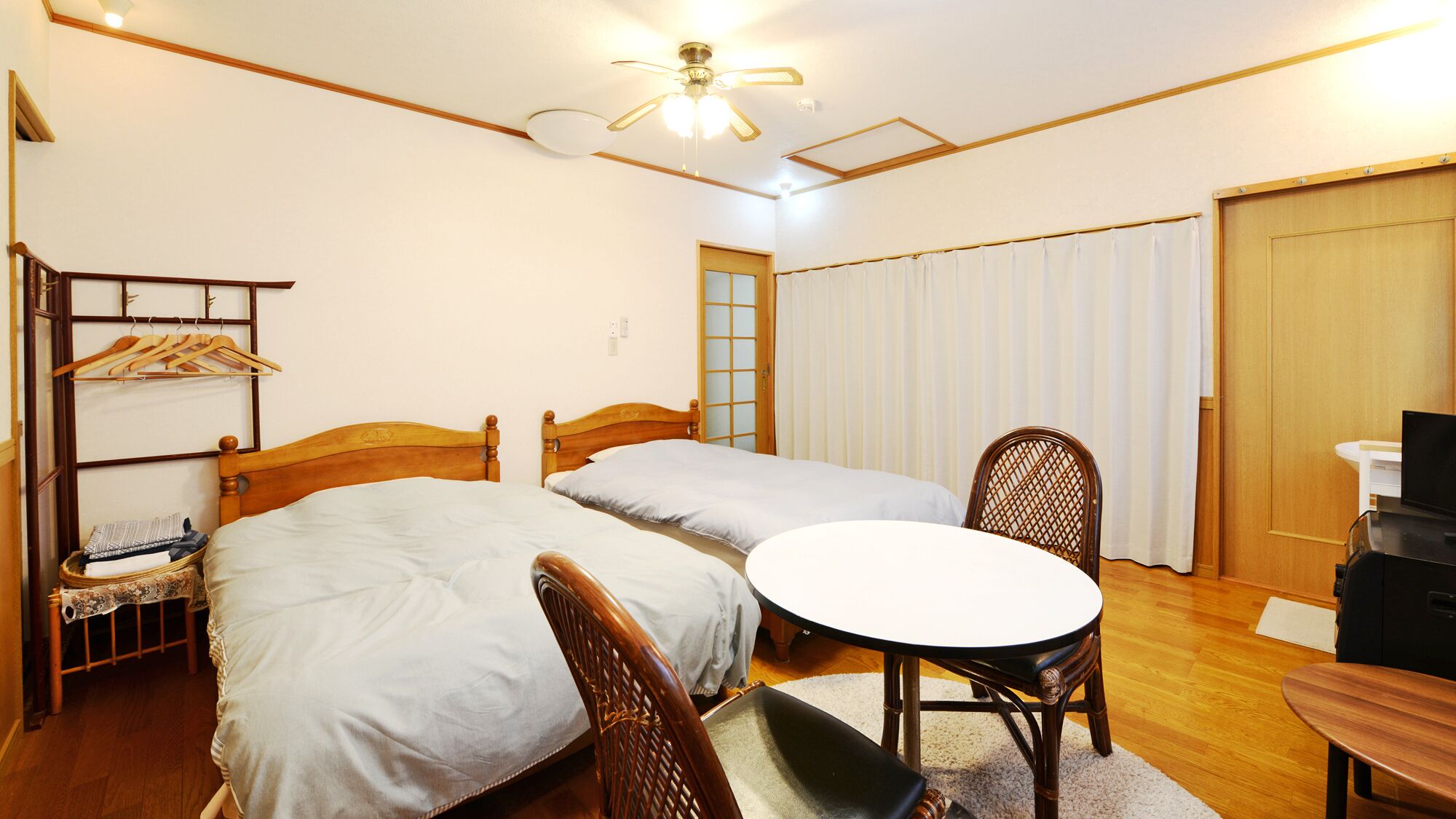 Habitación de hotel con dos camas individuales, una silla de madera, una mesita, un televisor y un ventilador de techo.