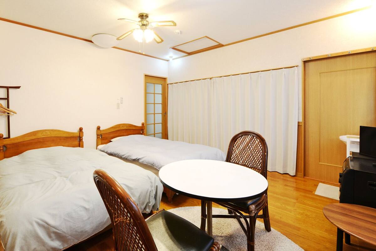 Habitación de hotel con dos camas individuales, una silla de madera, una mesita, un televisor y un ventilador de techo.