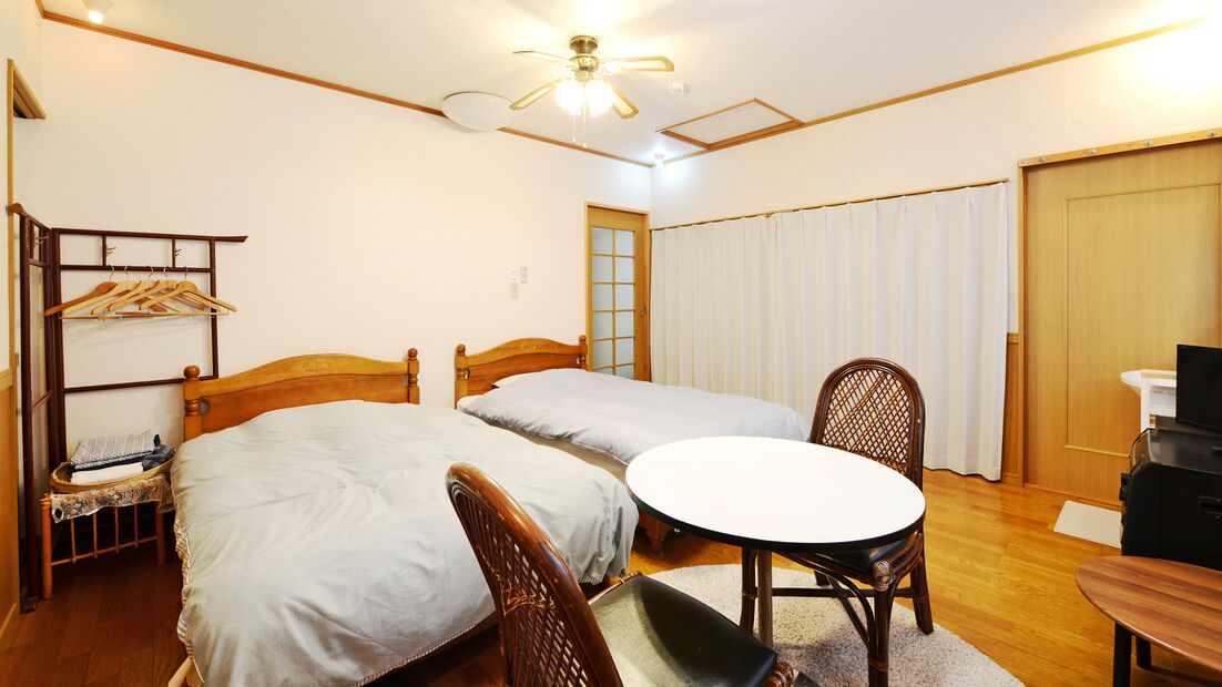 Une chambre d’hôtel avec deux lits simples, une table ronde, une chaise, une chaise en bois, une armoire et un ventilateur de plafond.