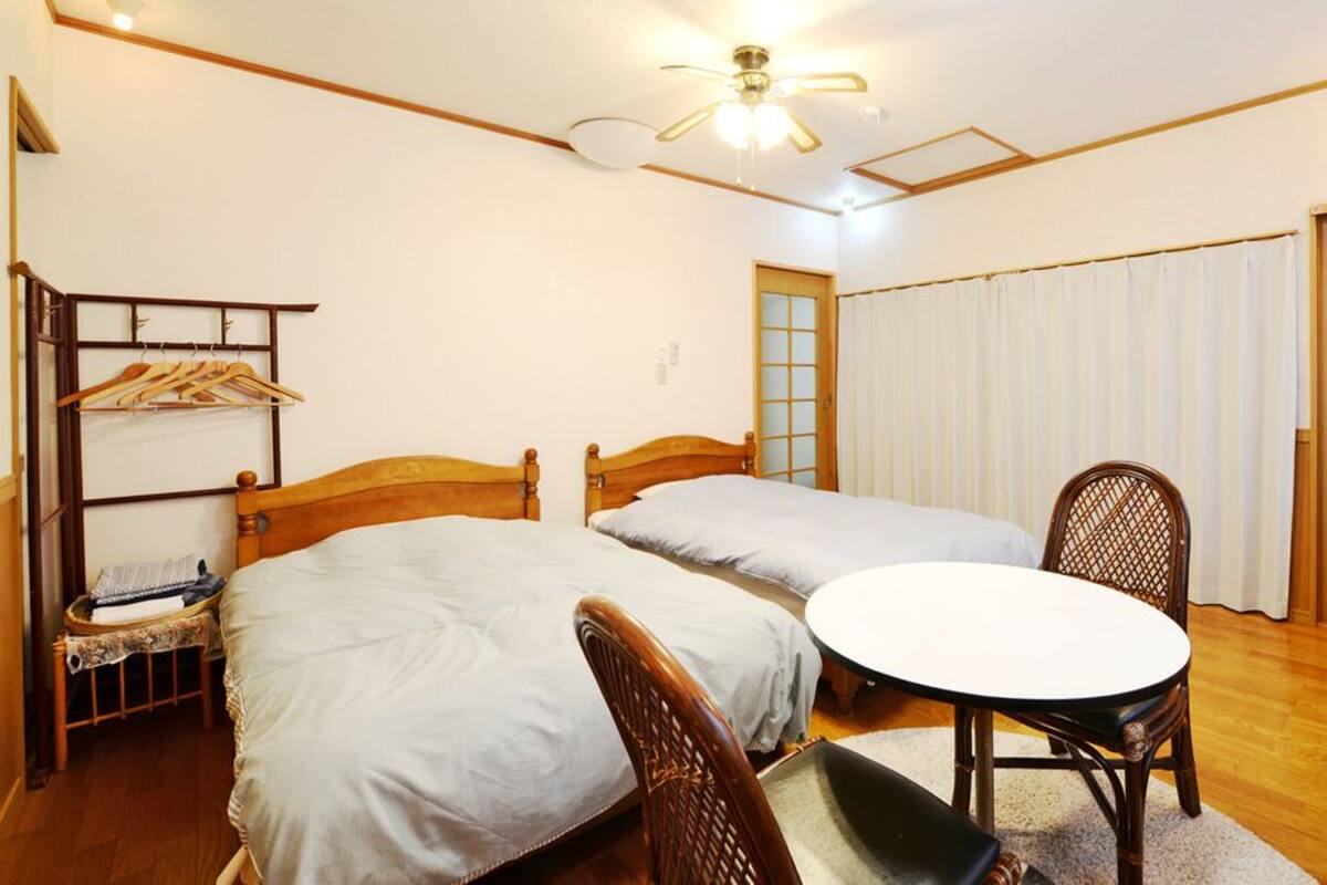 Habitación de hotel con dos camas individuales, una mesa redonda, una silla, un sillón de madera, un armario y un ventilador de techo.