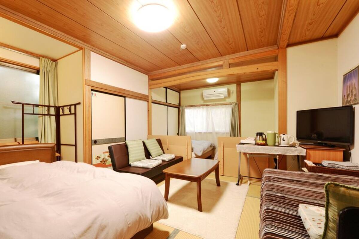 Una habitación con cama, sofá, una mesita, televisor y ventana con cortinas.