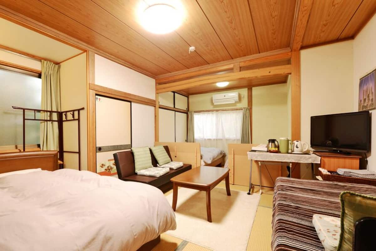 Una habitación con cama, sofá, una mesita, televisor y ventana con cortinas.