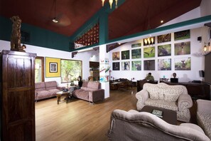 Un hall d’entrée spacieux avec un plafond haut, un plancher en bois et divers types de sièges, notamment des canapés et des fauteuils. Les murs sont ornés de tableaux représentant des animaux, et une grande fenêtre laisse entrer la lumière naturelle.