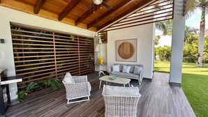 Un patio couvert doté d’un plafond en bois, d’un canapé gris, de deux fauteuils en osier, d’une petite table et d’un élément décoratif mural.
