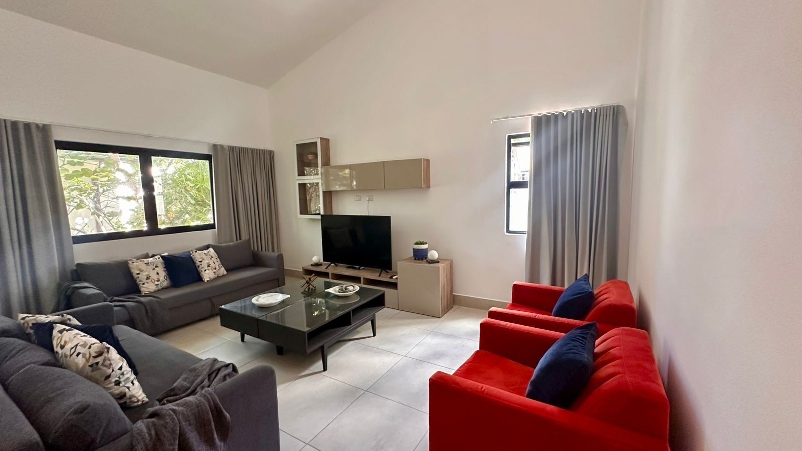 Un salon moderne avec un canapé gris, des fauteuils rouges, une table basse en verre, un téléviseur à écran plat et une petite table d’appoint.