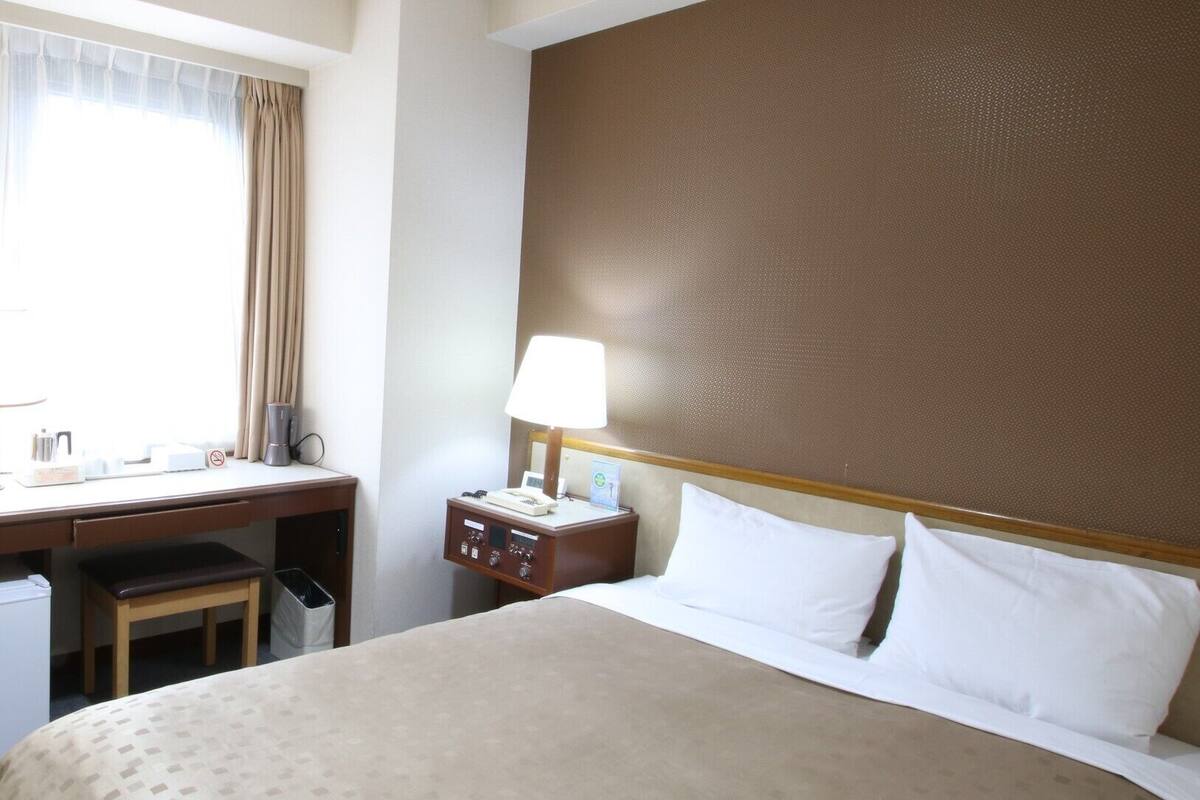 A hotel room with a bed, a desk, a chair, a nightstand with a lamp, and a window with curtains.