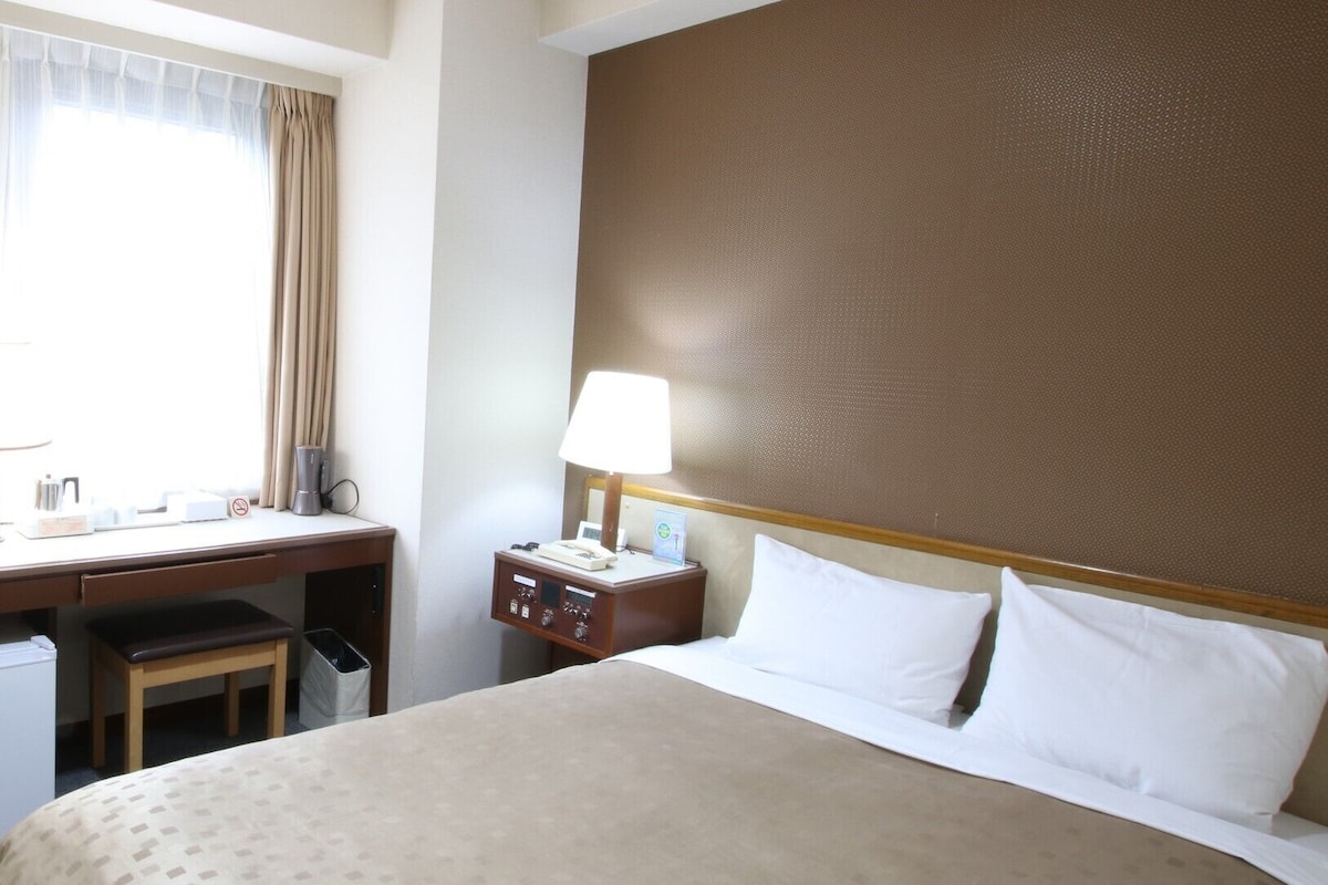 Une chambre d’hôtel comprenant un lit, un bureau, une chaise, une table de chevet avec une lampe et une fenêtre avec des rideaux.