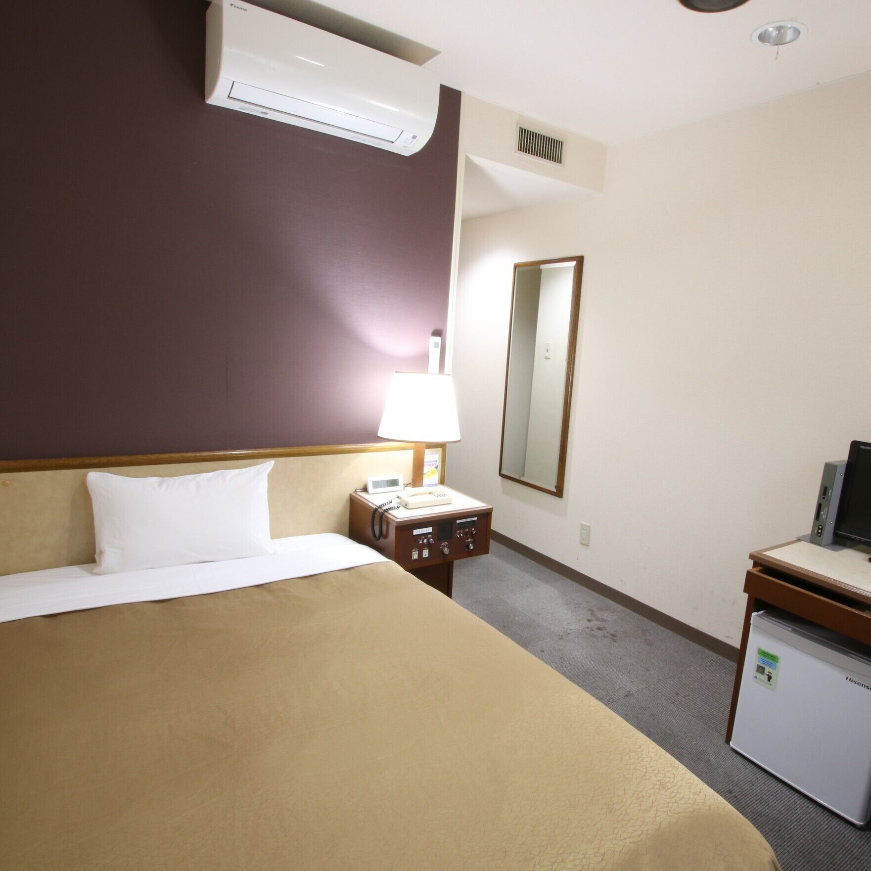 A hotel room with a bed, a desk with a computer, a phone, and a small refrigerator.