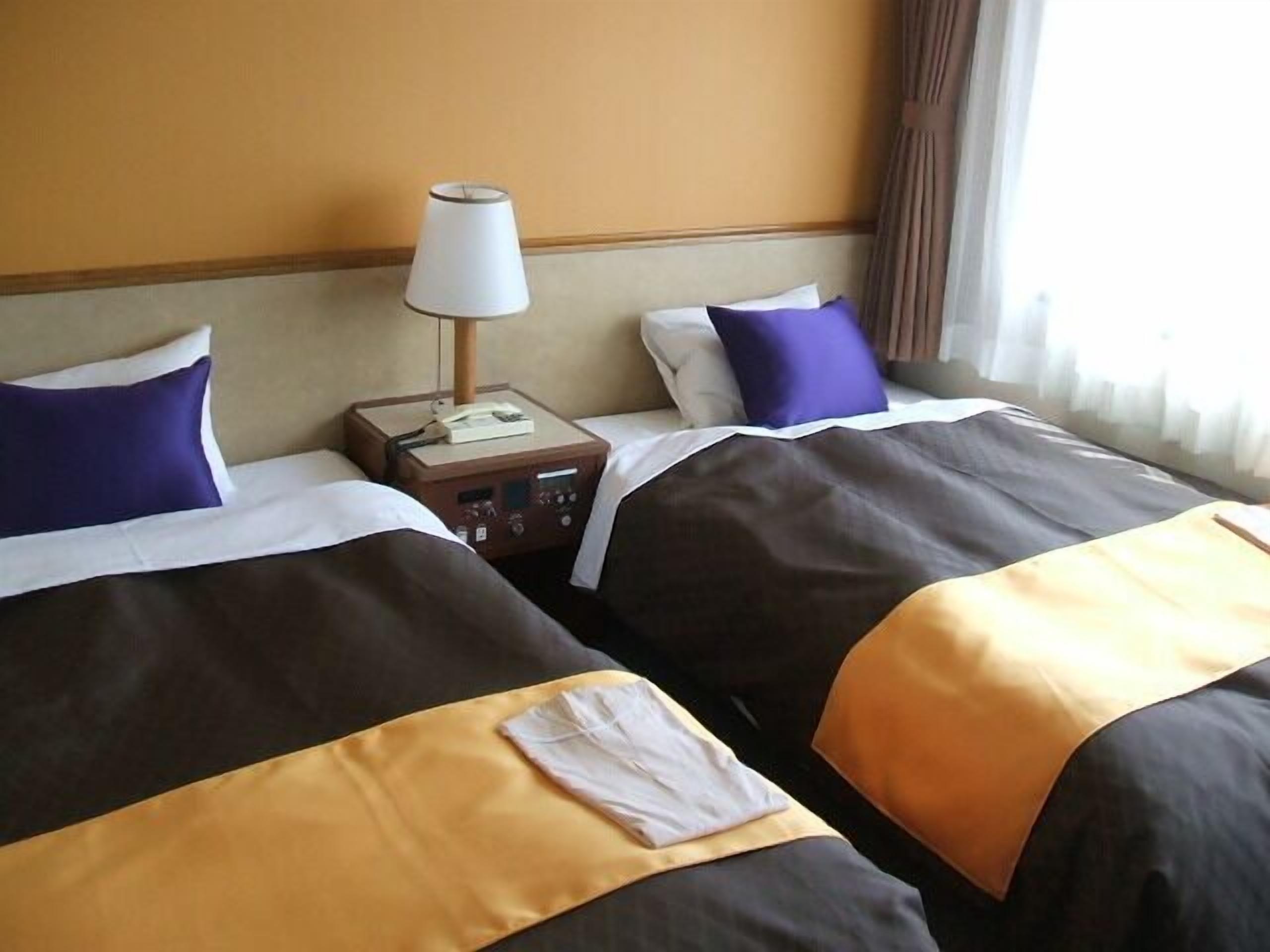 A hotel room with two beds, a nightstand with a lamp, and a window with curtains.