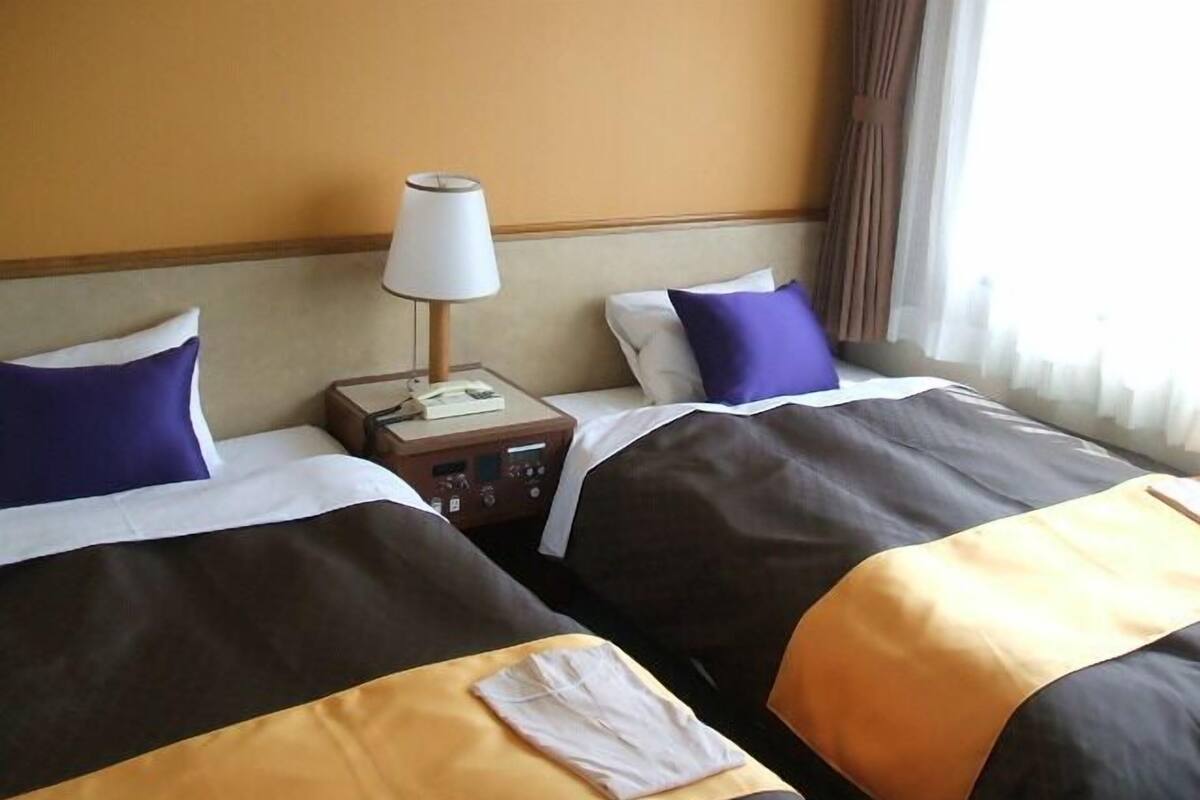 A hotel room with two beds, a nightstand with a lamp, and a window with curtains.