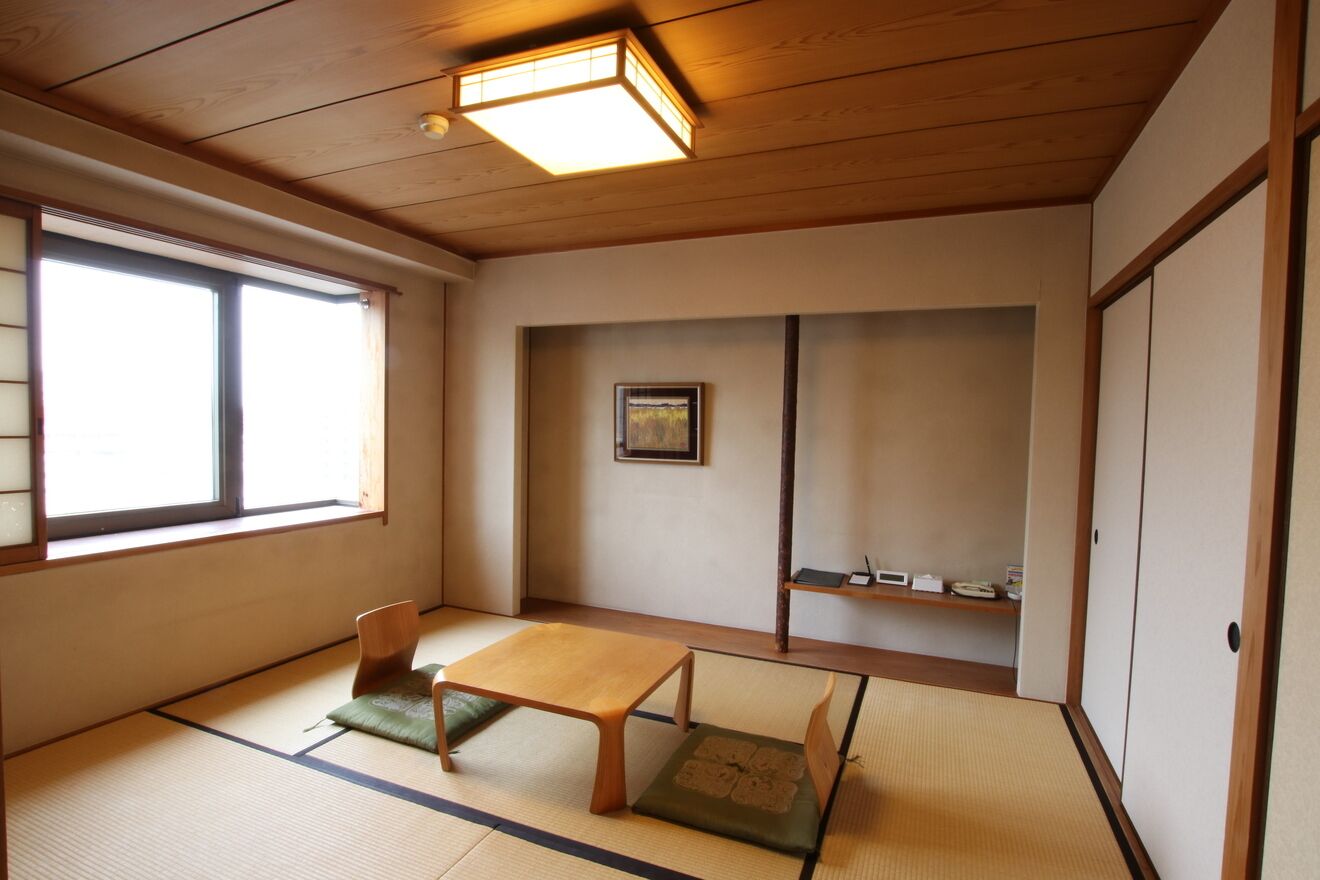 A traditional Japanese room with tatami flooring, a low wooden table, a chair, a window, and a shelf with items.