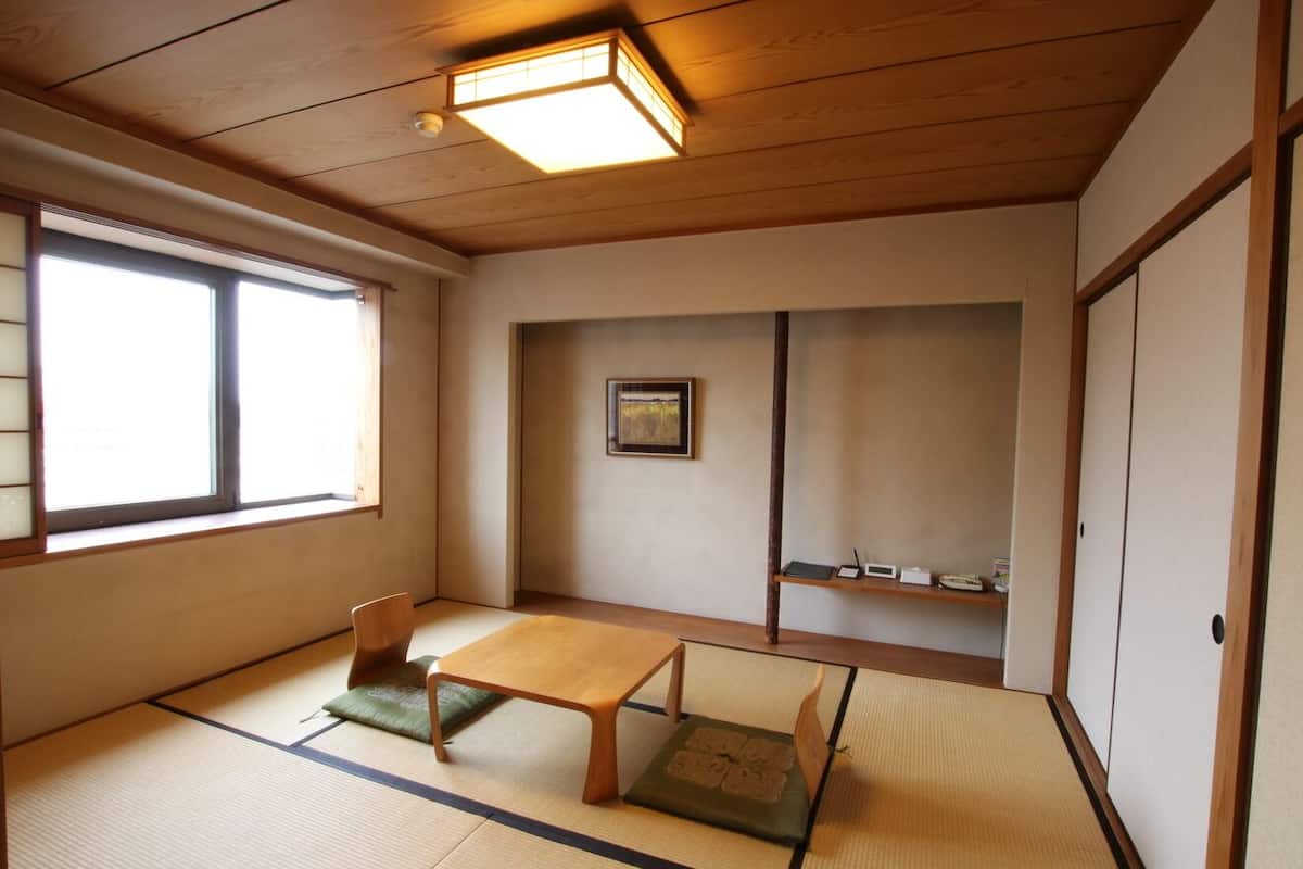 Une pièce de style japonais traditionnel, avec un sol recouvert de tatamis, une petite table en bois, une chaise, une fenêtre et une étagère où sont posés divers objets.