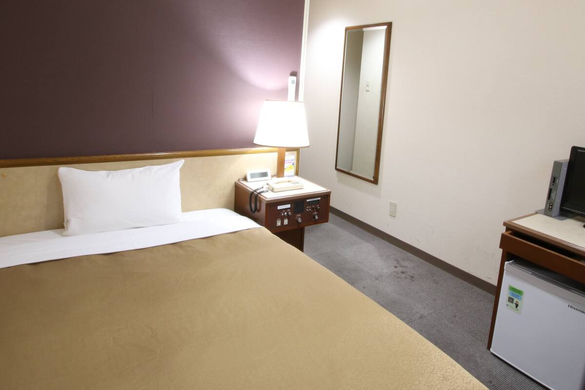 A hotel room with a bed, a desk with a computer, a lamp, a phone, and an air conditioning unit.