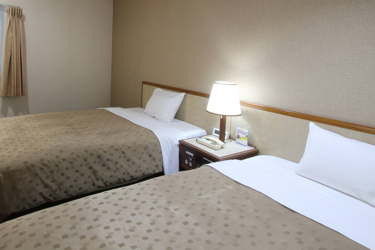 A hotel room with two beds, a nightstand with a lamp, a telephone, and a wall-mounted clock.