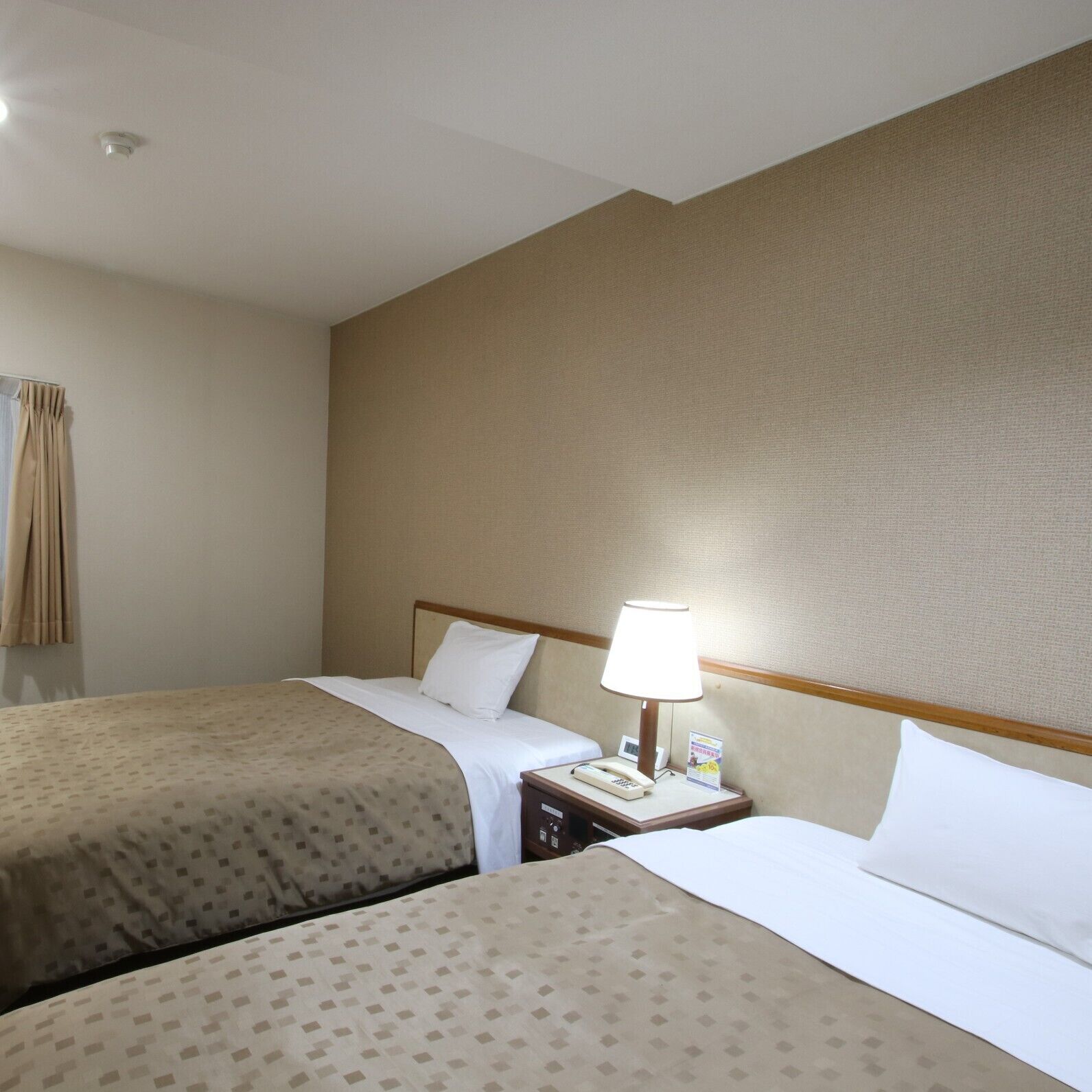 A hotel room with two beds, a bedside table with a lamp, a telephone, and a wall-mounted light fixture.