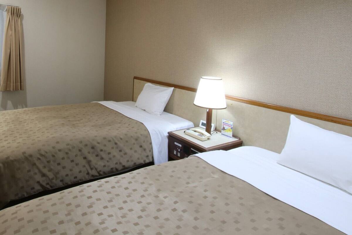 A hotel room with two beds, a bedside table with a lamp, a telephone, and a wall-mounted light fixture.
