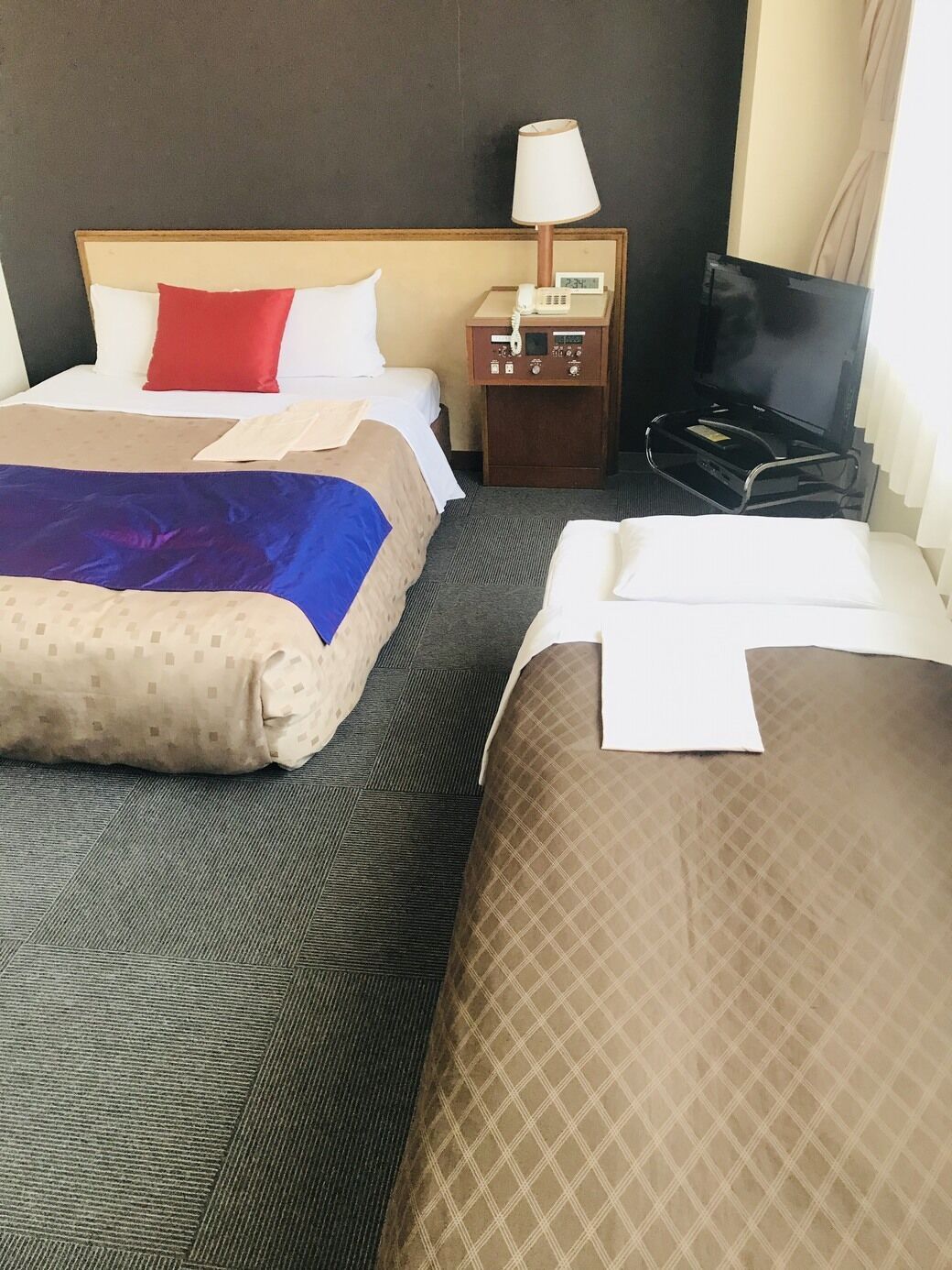 A hotel room with two beds, a nightstand with a lamp, a TV, and a clock.
