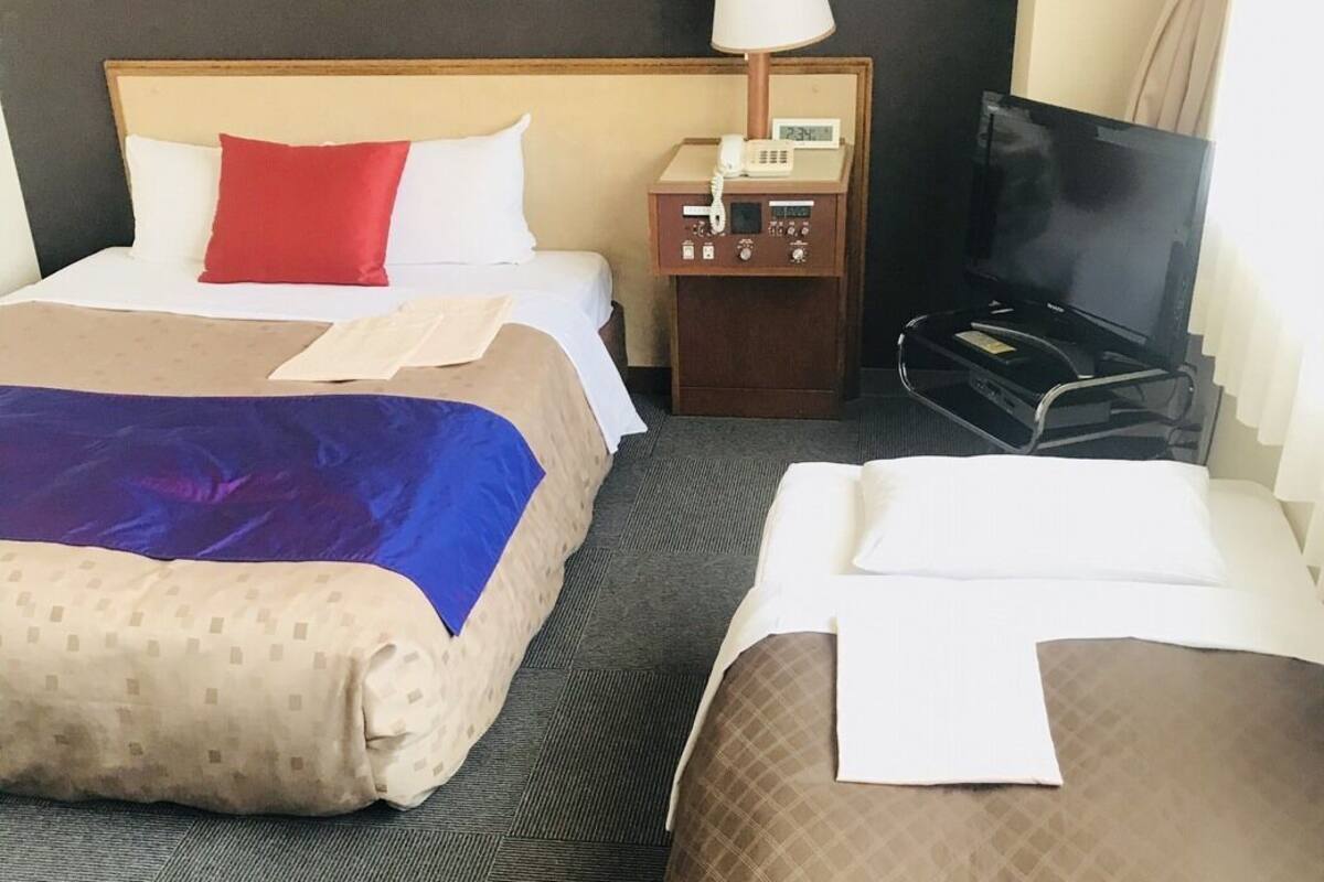 A hotel room with two beds, a nightstand with a lamp, a TV, and a clock.