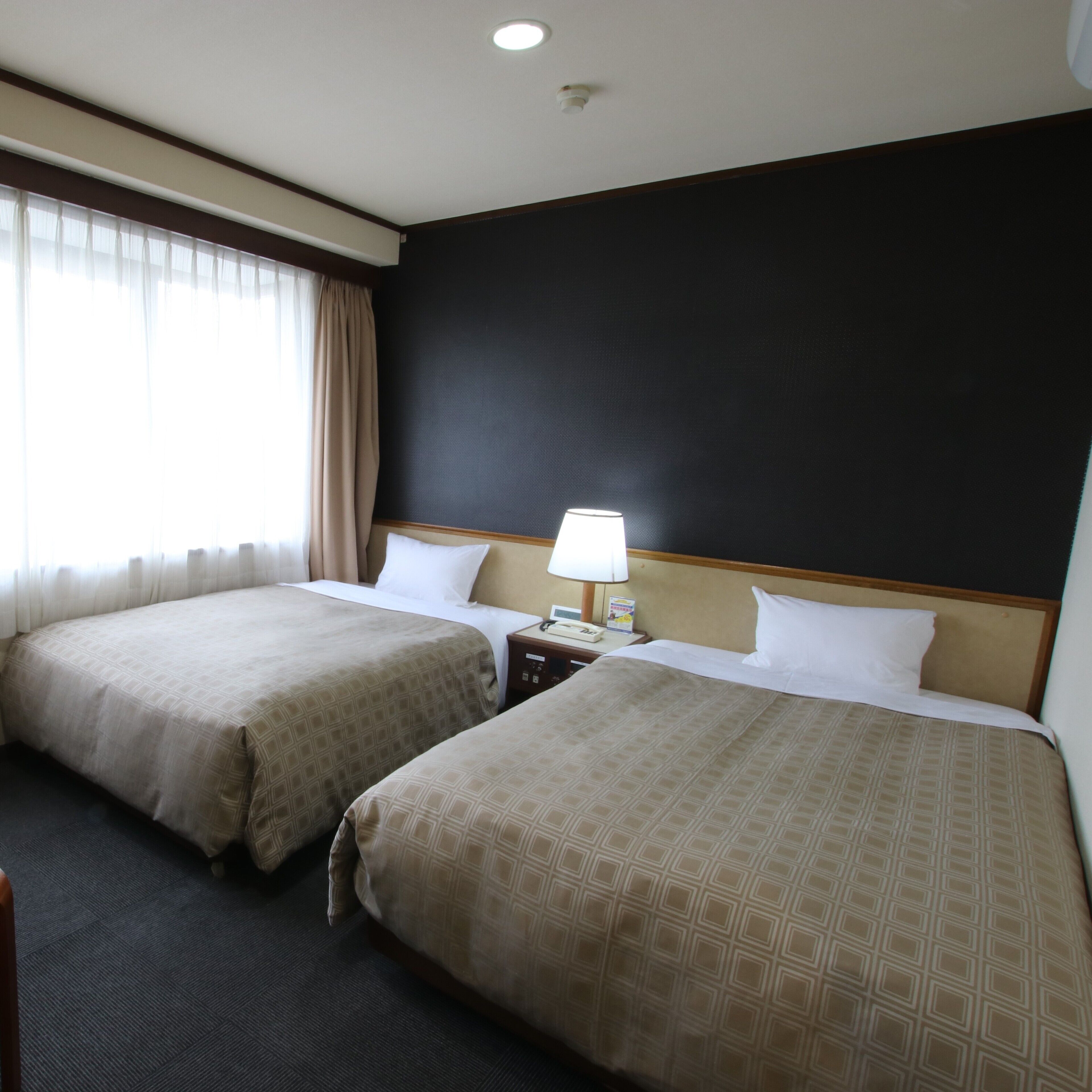 A hotel room with two beds, a nightstand with a lamp, and a window with curtains.