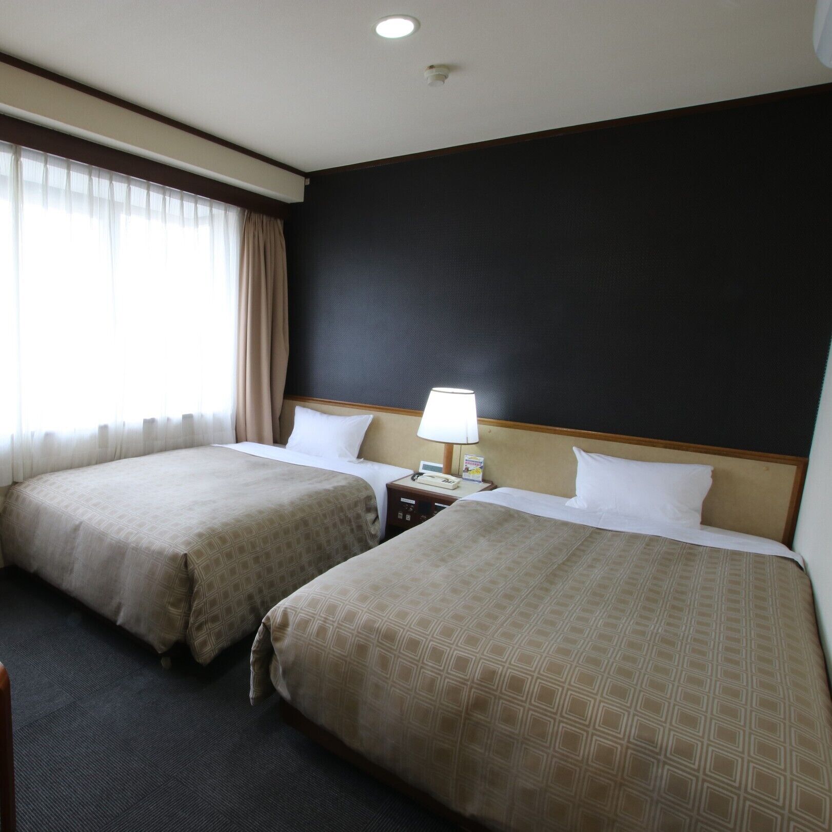 A hotel room with two beds, a nightstand with a lamp, and a window with curtains.