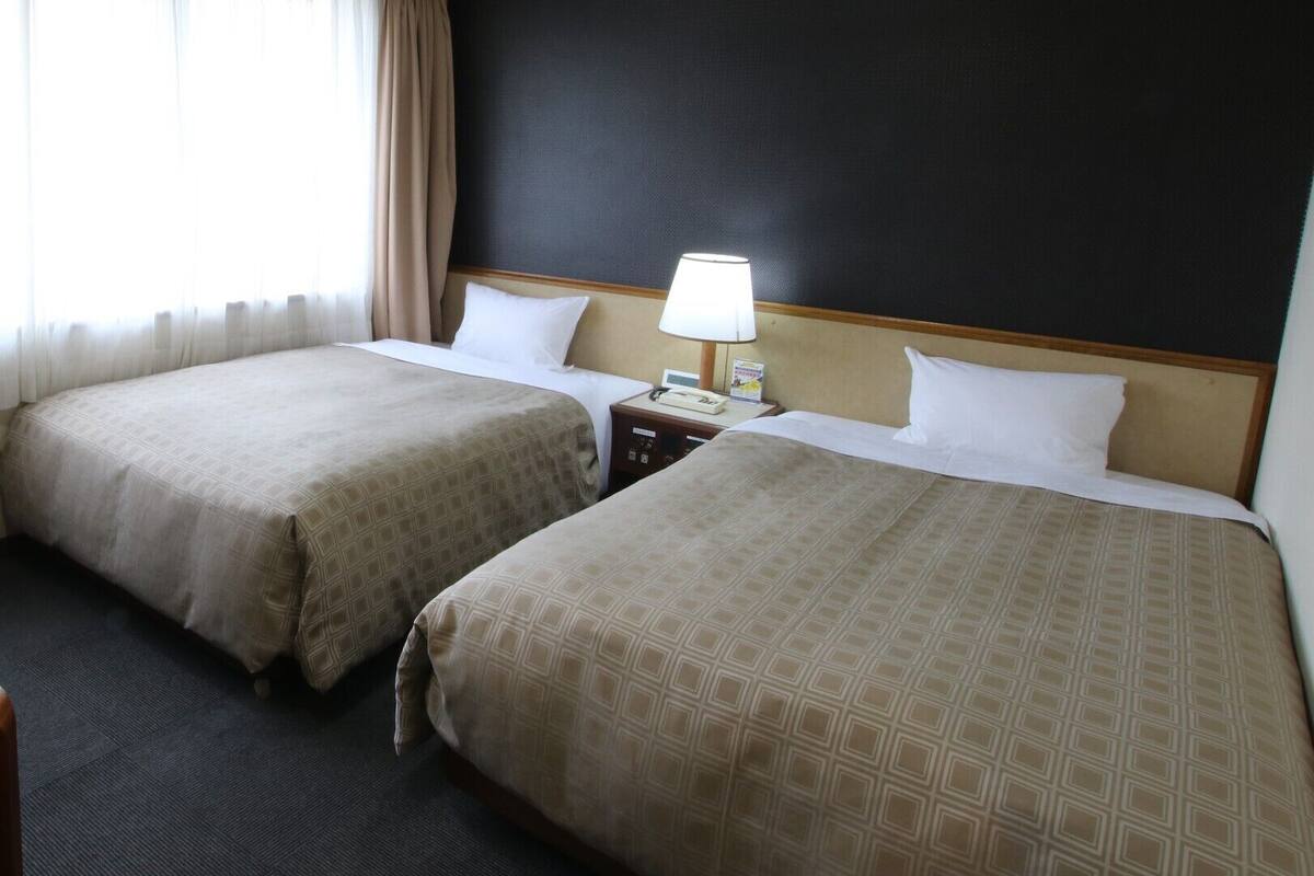 A hotel room with two beds, a nightstand with a lamp, and a window with curtains.