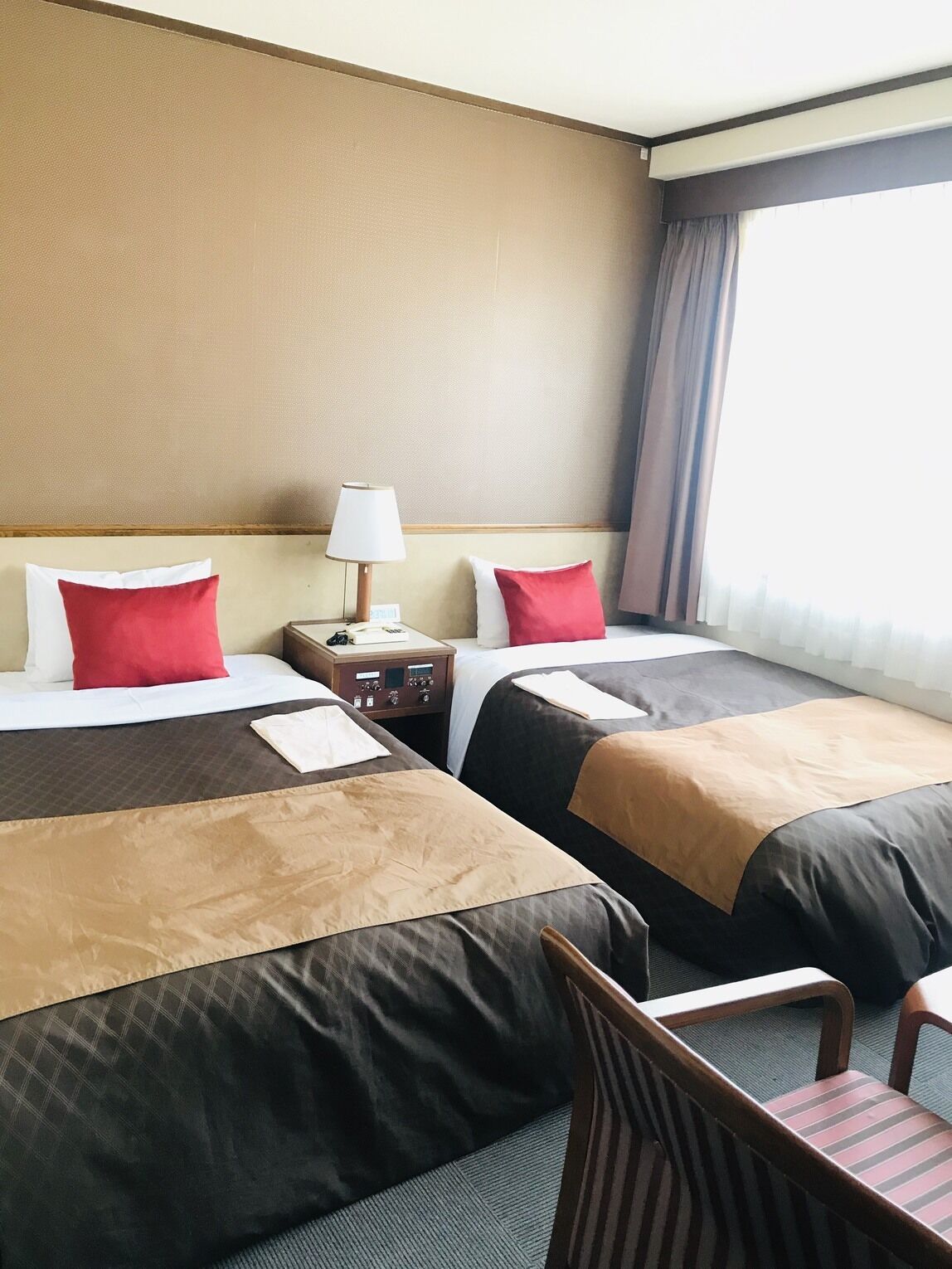 A hotel room with two beds, a nightstand with a lamp, a chair, and a view of the city through the window.