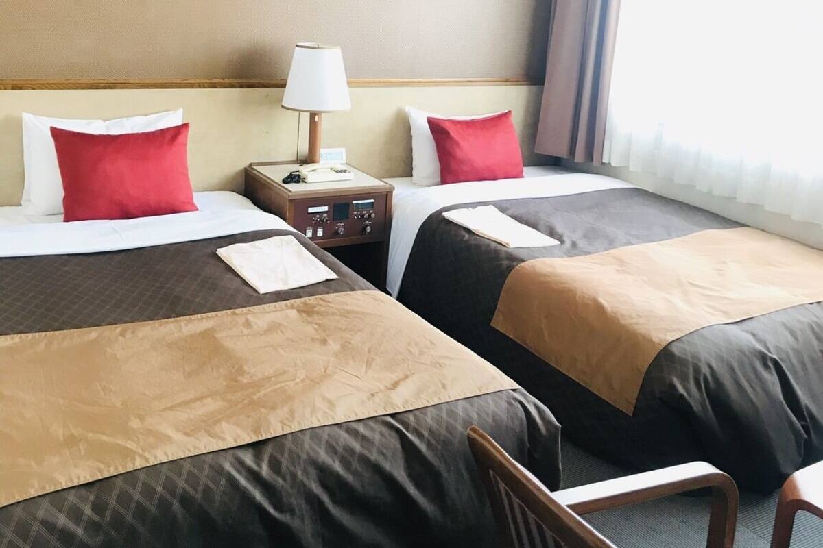 A hotel room with two beds, a nightstand with a lamp, a chair, and a view of the city through the window.