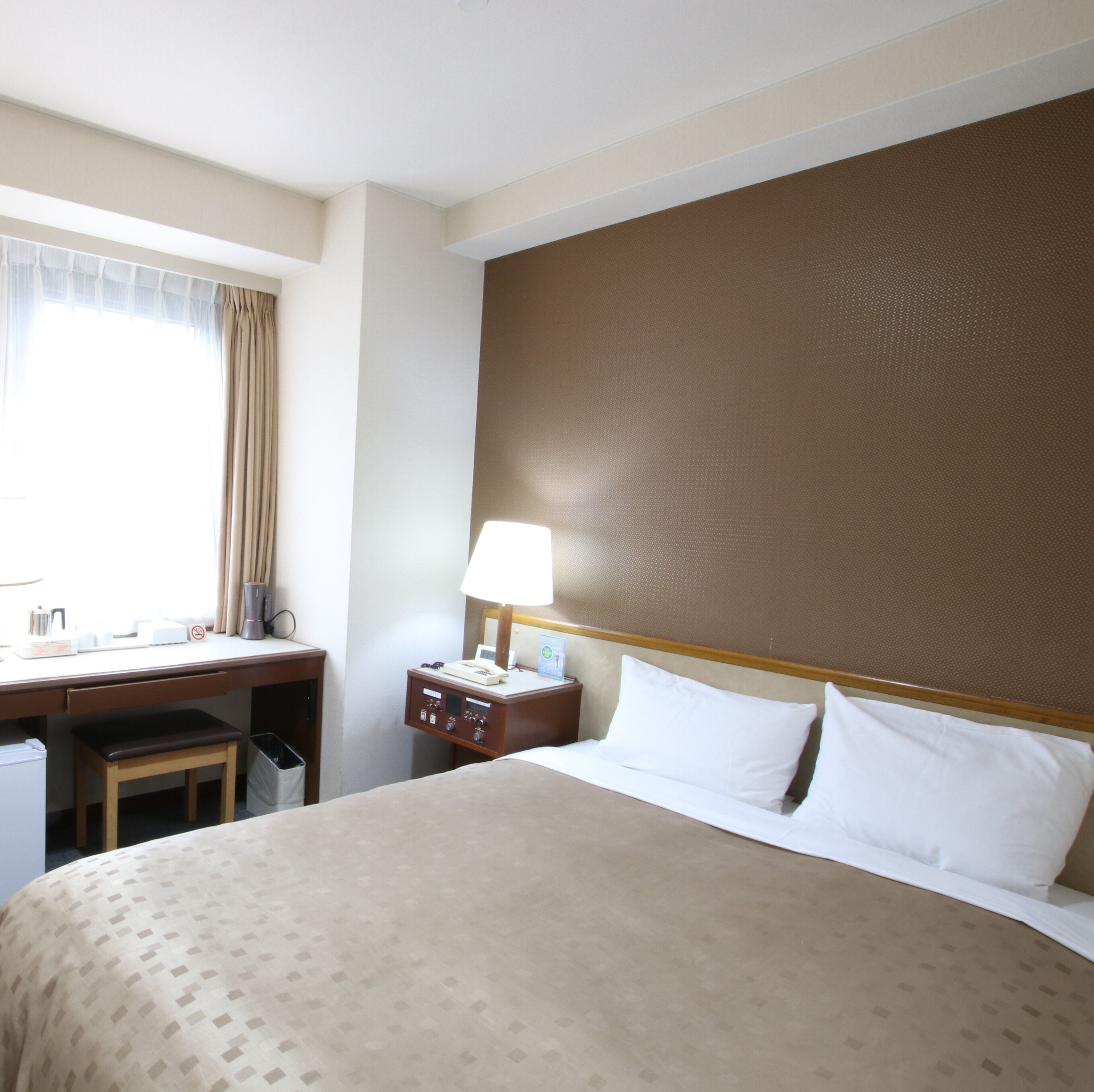 A hotel room with a bed, desk, chair, and a nightstand with a lamp.