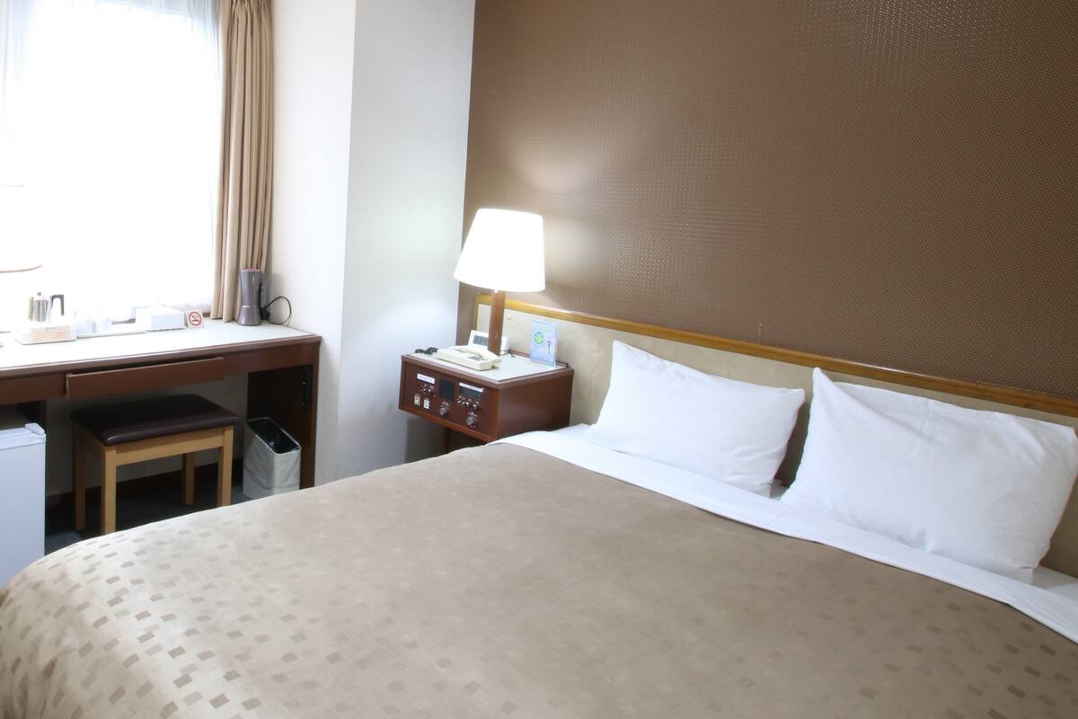A hotel room with a bed, desk, chair, and a nightstand with a lamp.
