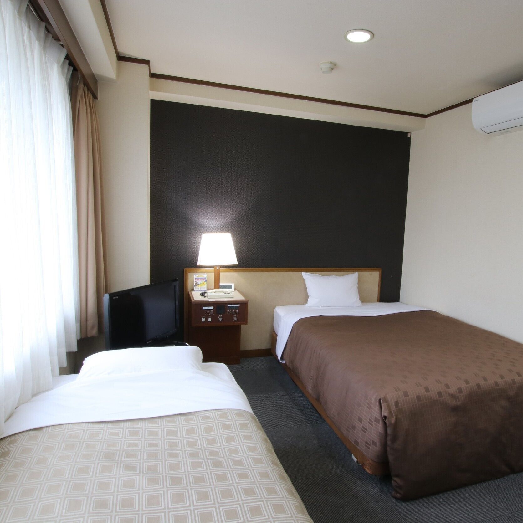 A hotel room with two beds, a desk with a lamp, a telephone, and a wall-mounted air conditioner.