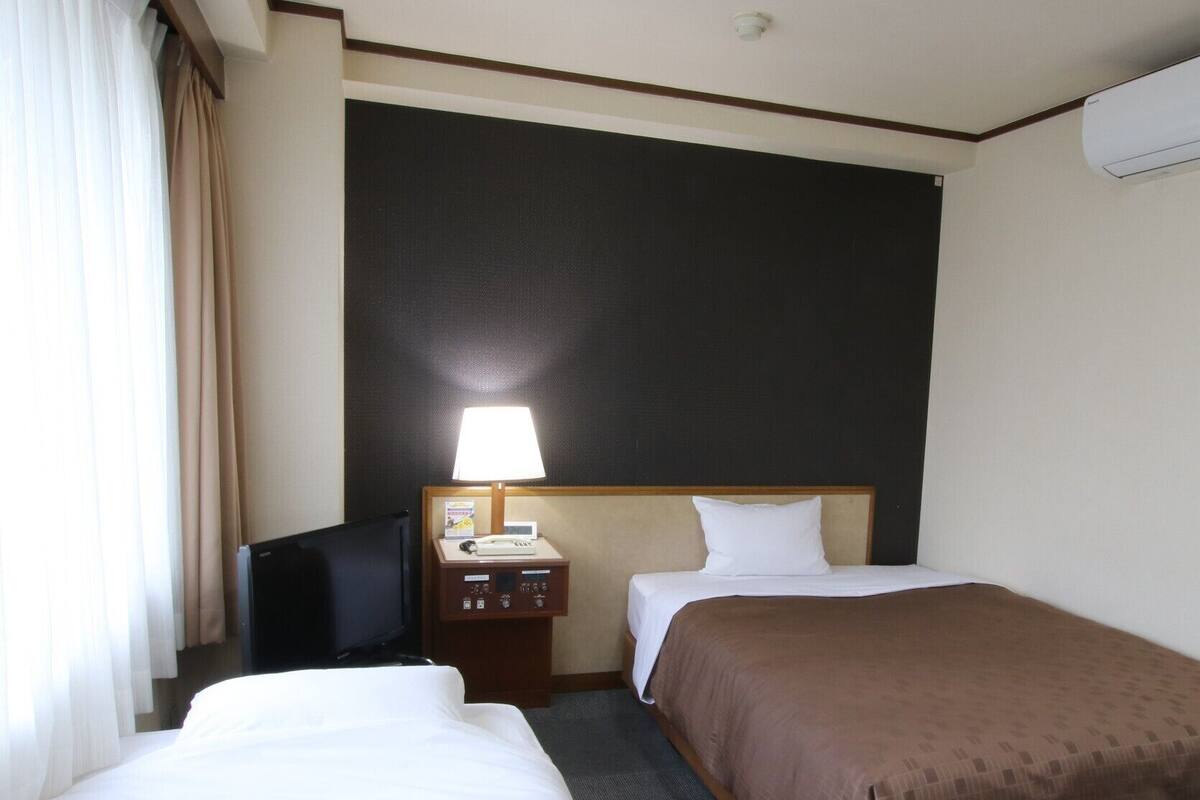 A hotel room with two beds, a desk with a lamp, a telephone, and a wall-mounted air conditioner.