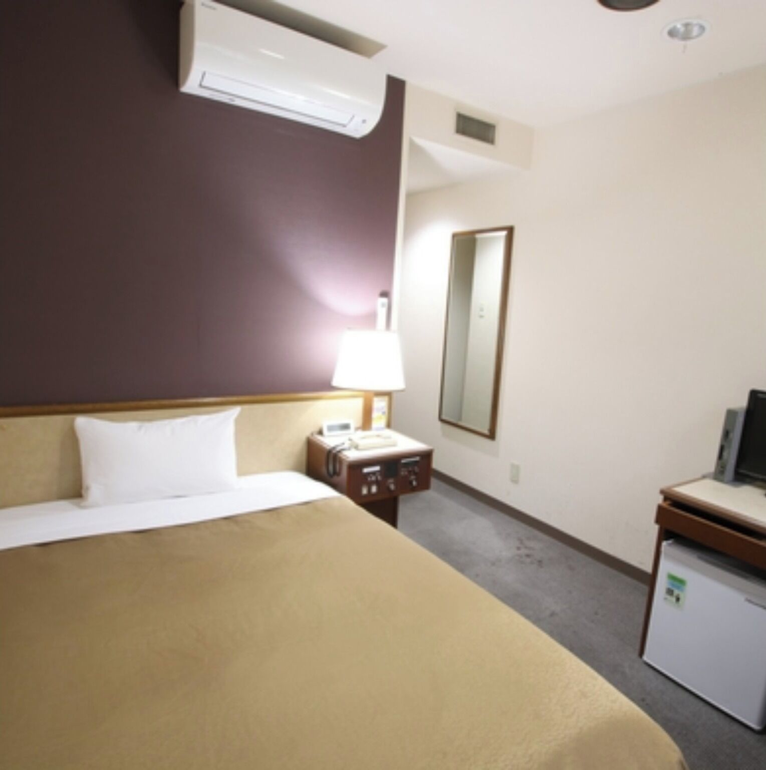A hotel room with a bed, a bedside table with a lamp, a small desk with a computer, and a mirror on the wall.