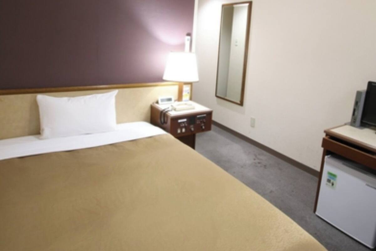 A hotel room with a bed, a bedside table with a lamp, a small desk with a computer, and a mirror on the wall.