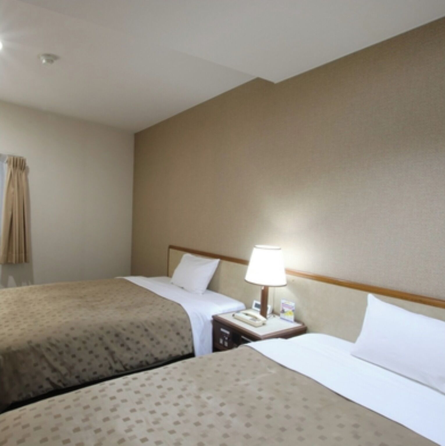 A hotel room with two beds, a nightstand with a lamp, and a window with curtains.