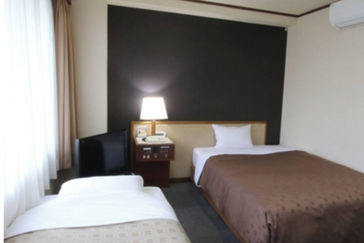 A hotel room with two beds, a desk with a lamp, a television, and a wall-mounted air conditioner.