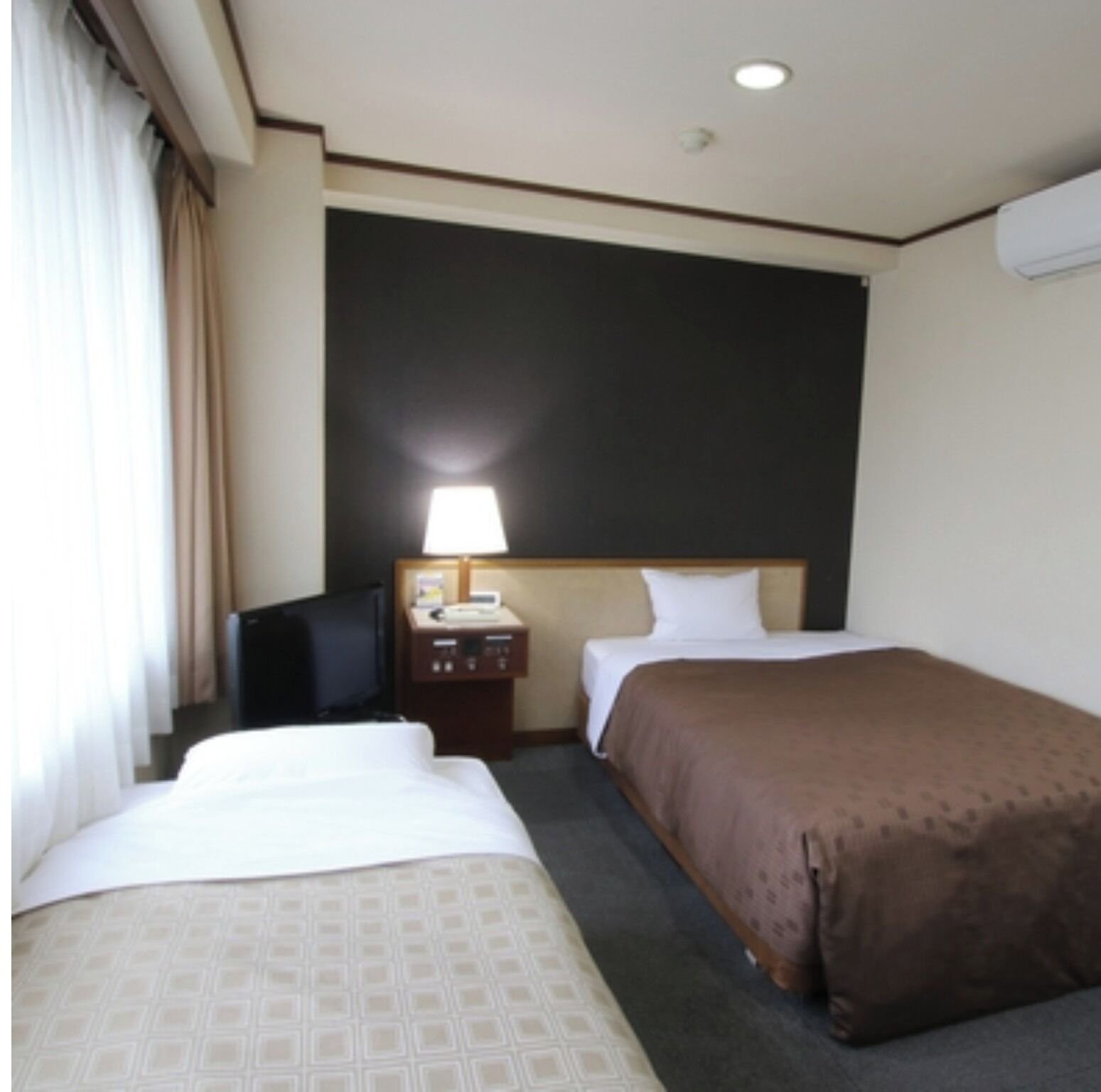 A hotel room with two beds, a desk with a lamp, a television, and a wall-mounted air conditioner.