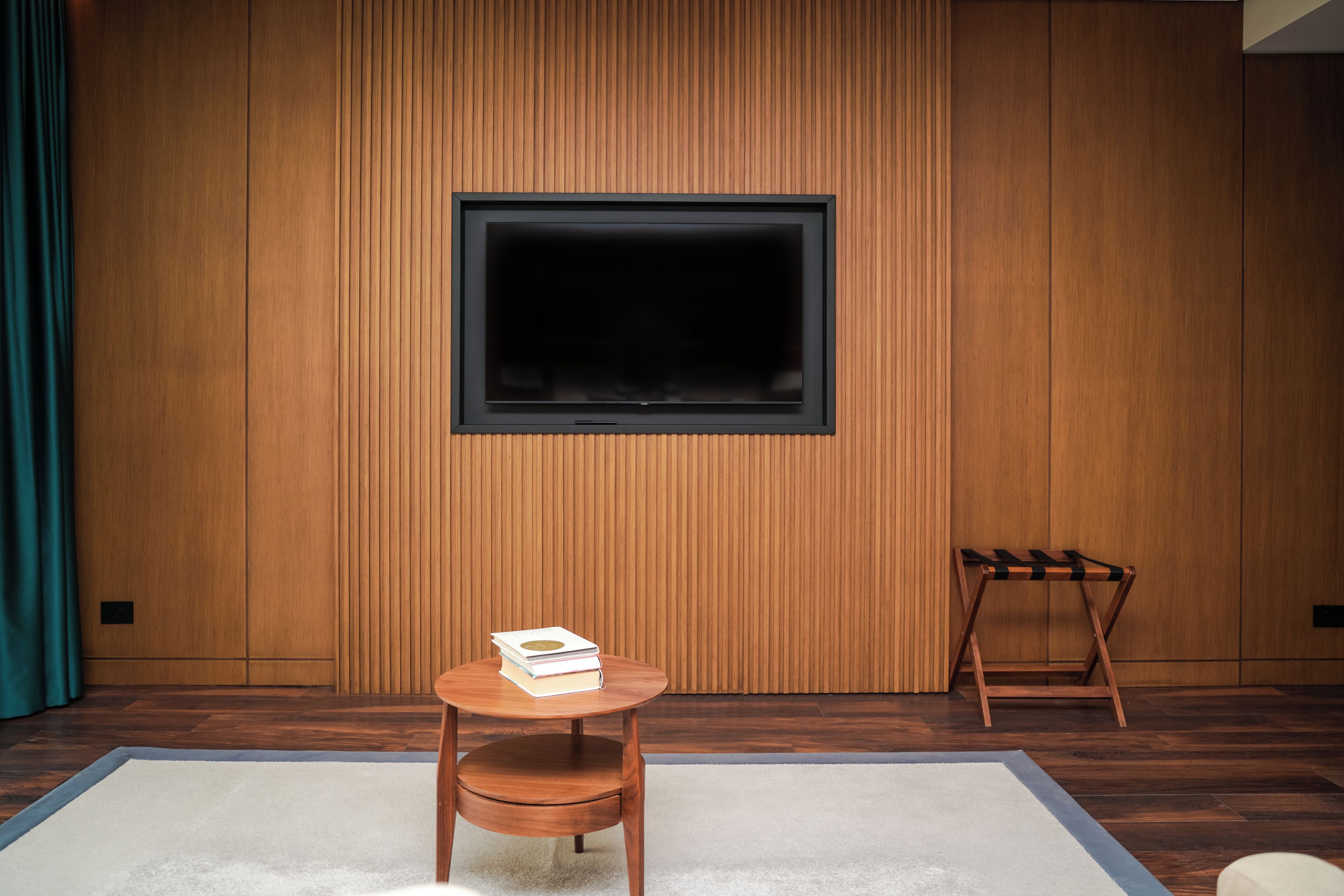 A modern room with a wooden paneled wall, a large TV mounted on it, a small round wooden table with a book, and a wooden chair.