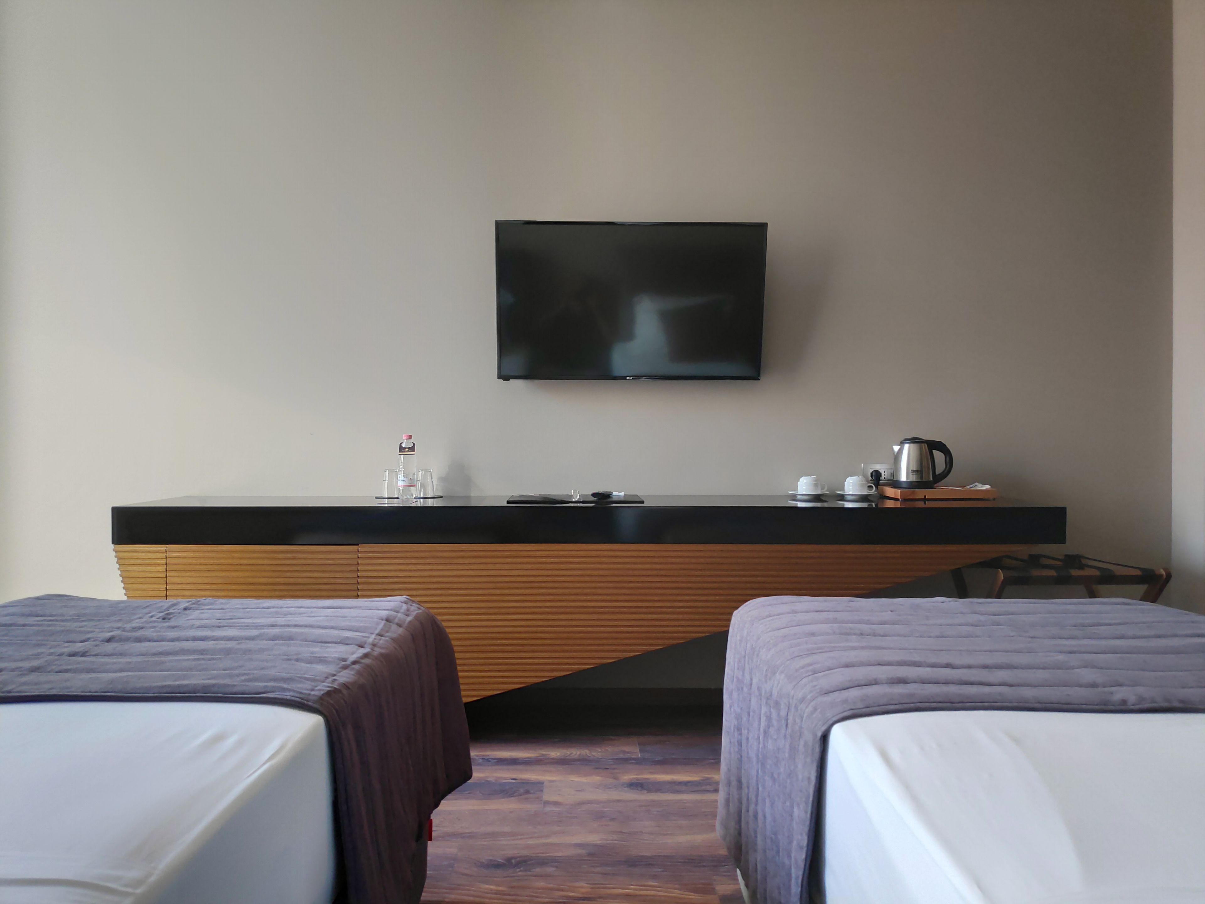 A hotel room with two beds, a flat-screen TV mounted on the wall, a small table with a kettle and cups, and a wooden desk with a bottle and glasses.