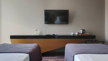 A hotel room with two beds, a flat-screen TV mounted on the wall, a small table with a kettle and cups, and a wooden desk with a bottle and glasses.