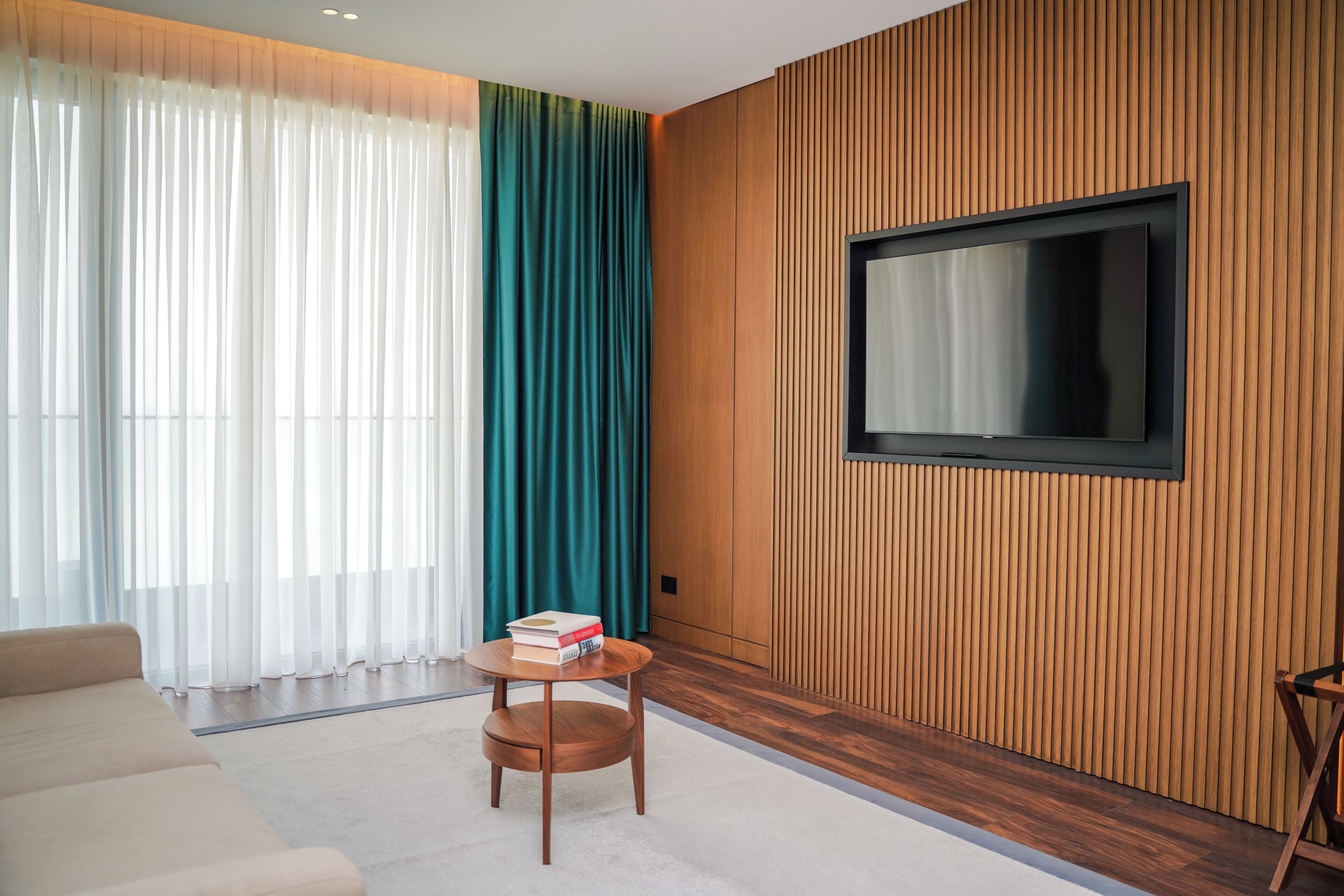 Una sala de estar moderna con un sofá, una pequeña mesa de madera con libros, un televisor montado en una pared de madera y cortinas translúcidas.
