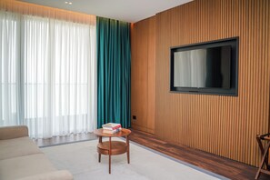 Una sala de estar moderna con un sofá, una pequeña mesa de madera con libros, un televisor montado en una pared de madera y cortinas translúcidas.