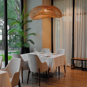 A dining area with a table set for two, surrounded by chairs, a hanging light fixture, and a potted plant. - Supreme Hotel (Golem)