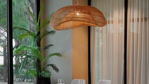 A dining area with a table set for two, surrounded by chairs, a hanging light fixture, and a potted plant. - Supreme Hotel (Golem)