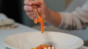 A person holding a piece of food with chopsticks over a white plate. - Supreme Hotel (Golem)