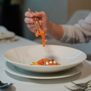 A person holding a piece of food with chopsticks over a white plate. - Supreme Hotel (Golem)